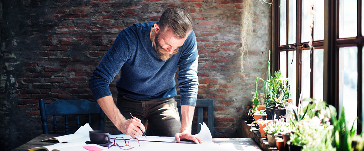 Ein Mann steht am Schreibtisch und zeichnet etwas mit einem Bleistift auf ein Blatt.