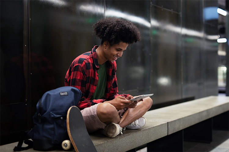 Ein Jugendlicher sitzt in einer U-Bahn auf der Ban und schaut auf ein Tablet. neben ihm steht ein Rucksack und ein Skateboard.