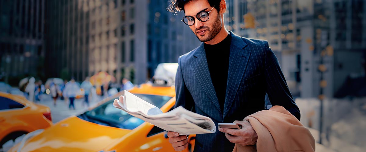 Mann liest Zeitung in der Öffentlichkeit, trinkt Kaffee und im Hintergrund steht ein gelbes Taxi.